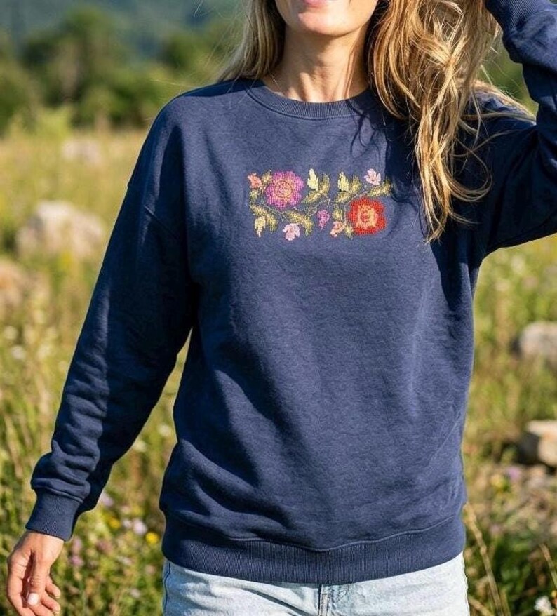 May include: A navy blue long-sleeve sweatshirt featuring a central embroidered design of two large flowers in shades of pink and red, surrounded by smaller green leaves and delicate floral accents, with a ribbed crew neckline and cuffs.