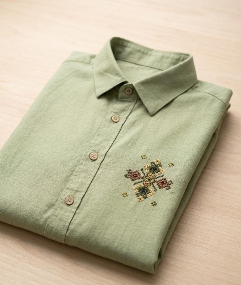 Puede incluir: Una camisa de color verde salvia claro con cuello en punta y tapeta central con botones a juego. La parte delantera de la camisa presenta un peque&ntilde;o y detallado patr&oacute;n de bordado en punto de cruz en tonos marr&oacute;n, verde y amarillo.