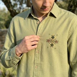 Puede incluir: Una camisa de manga larga con botones en color verde oliva claro. La camisa presenta un cuello en punta y una hilera vertical central de botones a juego. En el pecho izquierdo, hay un peque&ntilde;o y complejo bordado geom&eacute;trico en punto de cruz.