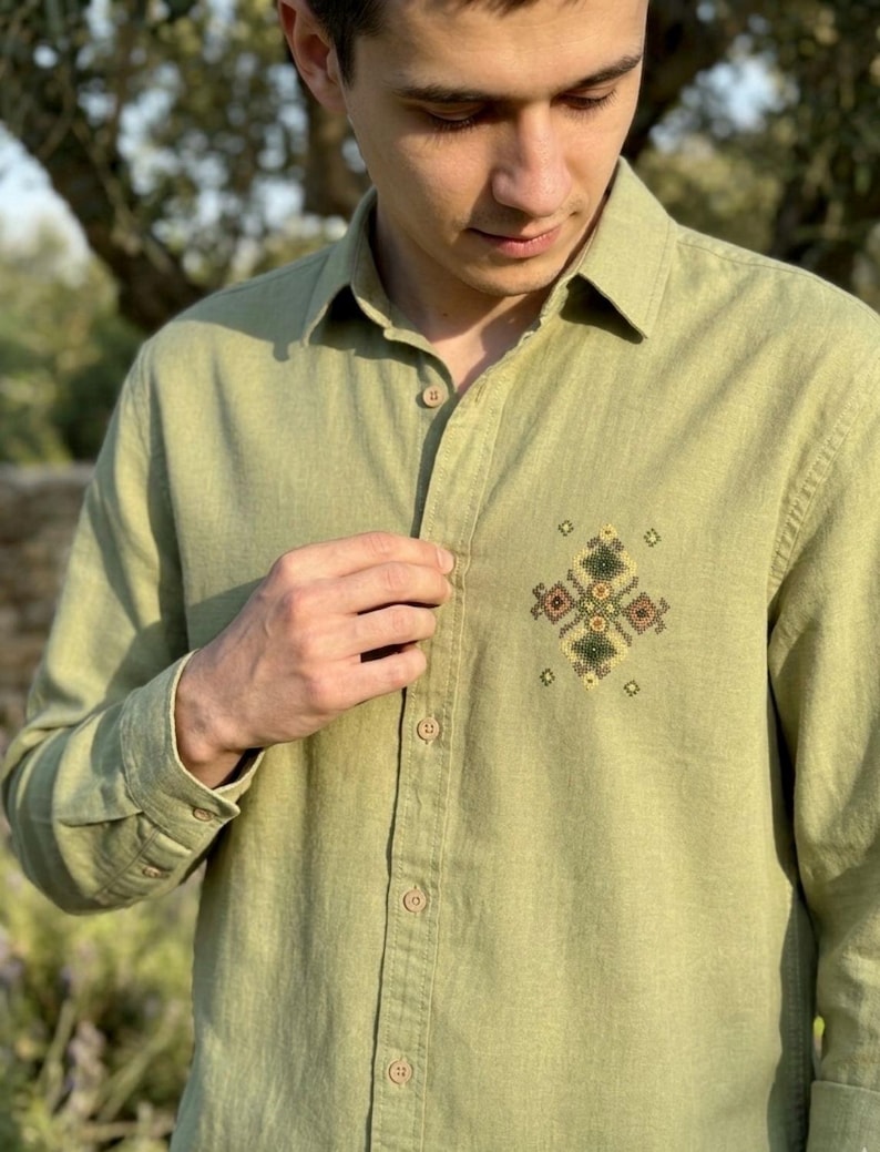 Puede incluir: Una camisa de manga larga con botones en color verde oliva claro. La camisa presenta un cuello en punta y una hilera vertical central de botones a juego. En el pecho izquierdo, hay un peque&ntilde;o y complejo bordado geom&eacute;trico en punto de cruz.