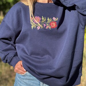 May include: A navy blue crewneck sweatshirt featuring a central cross-stitch embroidery design of various flowers in shades of red, pink, orange, and green. The garment has long sleeves with ribbed cuffs and a ribbed neckline, paired with light blue denim jeans.