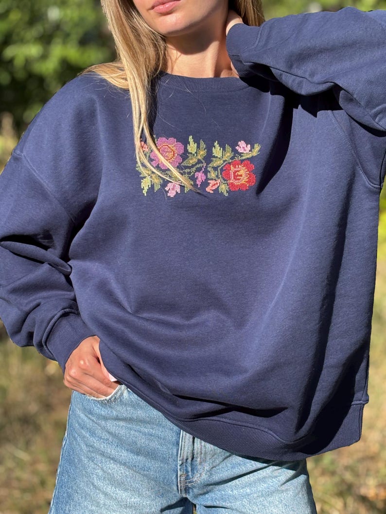 May include: A navy blue crewneck sweatshirt featuring a central cross-stitch embroidery design of various flowers in shades of red, pink, orange, and green. The garment has long sleeves with ribbed cuffs and a ribbed neckline, paired with light blue denim jeans.