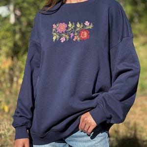 May include: A navy blue crewneck sweatshirt featuring a central embroidered design of colourful flowers and green leaves. The long-sleeved garment has a relaxed fit with ribbed cuffs and a ribbed neckline, paired with light blue denim shorts.