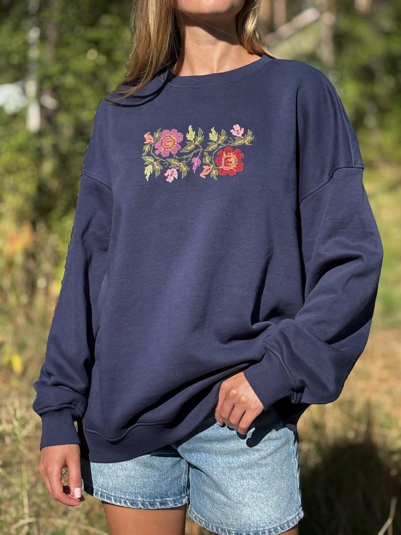May include: A navy blue crewneck sweatshirt featuring a central embroidered design of colourful flowers and green leaves. The long-sleeved garment has a relaxed fit with ribbed cuffs and a ribbed neckline, paired with light blue denim shorts.