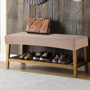 Puede incluir: Un banco tapizado beige con estructura de madera y una balda inferior con varios pares de zapatos. Un bolso tote de cuero marrón está encima. El banco está contra una pared de madera.