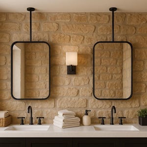 May include: Bathroom vanity with two black-framed mirrors suspended from the ceiling. The mirrors are above two sinks with black faucets. A wall-mounted light fixture and a stone brick wall complete the design. A basket with towels and a small plant are also visible.