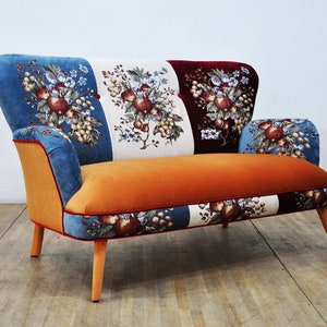 May include: A small, orange, two-seater sofa with a patchwork design. The fabric features a floral pattern with red, blue, and white accents. The sofa has wooden legs.