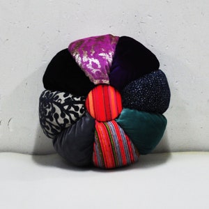 May include: A round, multi-coloured, flower-shaped cushion with a red and orange striped centre. The cushion is made of different fabrics, including velvet, patterned fabric, and brocade.