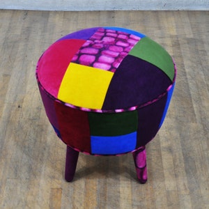 Può includere: Un pouf rotondo in patchwork multicolore con tre gambe in legno. Il pouf è rivestito con una varietà di tessuti, tra cui rosa, viola, giallo, verde e blu.