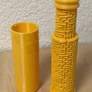 May include: A yellow 3D-printed puzzle toy with a cylindrical design. One piece is a simple cylinder, and the other is a taller cylinder with a maze pattern. The toy is designed to be a brain teaser.