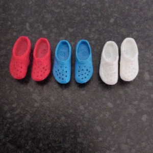 May include: Three pairs of miniature clogs in red, blue, and white. Each pair has a rounded toe and several small holes. The clogs are made of a solid material and are arranged in a row against a dark background.