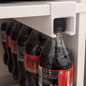 以下が含まれることがあります： 白いプラスチック製の棚オーガナイザーは、いくつかのコカ・コーラ ゼロシュガーのボトルを保持しています。ボトルは黒色で赤いラベルとコカ・コーラのロゴが付いています。オーガナイザーは白い棚に取り付けられています。