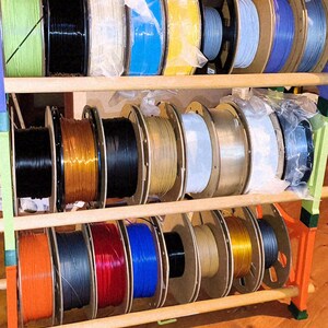 May include: A wooden rack displaying multiple spools of 3D printer filament in various colors, including green, black, white, blue, yellow, and gray. The spools are neatly arranged on multiple shelves, showcasing a diverse selection of materials for 3D printing projects.