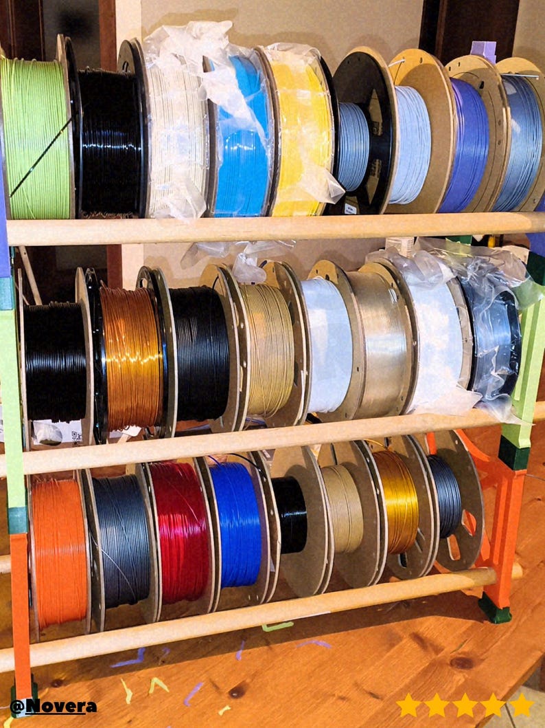 May include: A wooden rack displaying multiple spools of 3D printer filament in various colors, including green, black, white, blue, yellow, and gray. The spools are neatly arranged on multiple shelves, showcasing a diverse selection of materials for 3D printing projects.
