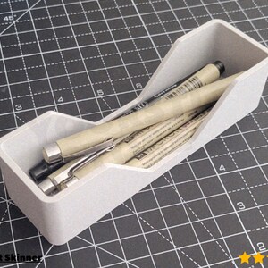 May include: A light grey pen holder with a stepped design, containing several pens. The pens are beige with silver accents. The holder is on a black cutting mat with a grid pattern. The image includes the text "@Robert Skinner".