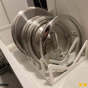 May include: A white metal pot lid organizer holding several stainless steel pot lids. The organizer is inside a white cabinet. The lids are of varying sizes and have metal handles. The organizer has multiple slots to hold the lids upright.