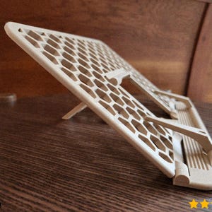 Peut inclure: Un support de tablette géométrique beige clair avec un motif en nid d'abeille. Le support est incliné et soutenu par un petit pied. Le support est en matériau de couleur claire et est conçu pour contenir une tablette ou un appareil similaire.
