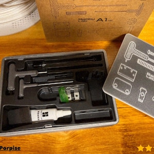 May include: A grey tool kit containing various tools, including T-wrenches, brushes, and a small bottle of adhesive. The kit is in a black tray, with a cardboard box and a grey plate with diagrams in the background. The text "Bambu Lab A1 mini" is visible.