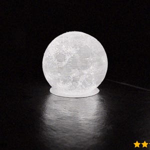 May include: A white, spherical moon lamp with a textured surface, resting on a small, circular base. The lamp is illuminated, casting a soft glow against a dark background. The lamp is a decorative lighting fixture.