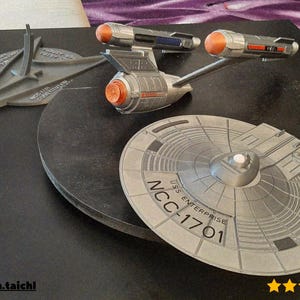 May include: A silver model of the USS Enterprise NCC-1701 starship, with orange and blue accents, and a stand. The model is on a black surface. The saucer section is visible, with the text "USS Enterprise NCC-1701".