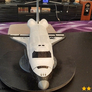 Puede incluir: Un modelo detallado de un transbordador espacial, predominantemente blanco con detalles en gris, se exhibe sobre una base circular negra. El modelo presenta detalles intrincados, incluyendo la marca USA y la forma icónica del transbordador. Es un artículo de colección.