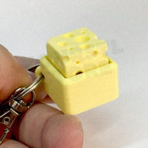 以下が含まれることがあります： 長方形のベースとチーズブロックが付いた、黄色のチーズ型キーホルダー。チーズブロックにはいくつかの穴があります。キーホルダーは、シルバーの金属リングと留め金に取り付けられています。「@FlexiPals Toy Shop」というテキストが表示されます。