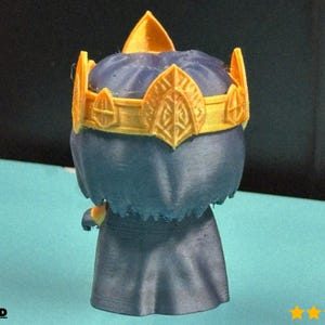 Puede incluir: Figura impresa en 3D de un personaje que lleva una corona dorada. La figura es principalmente azul oscuro con una corona dorada. La corona tiene detalles intrincados y una parte superior puntiaguda. La figura está sobre una superficie azul claro.