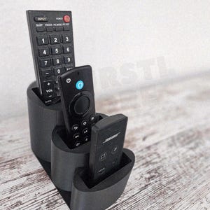 May include: A black 3-slot remote control holder with three remotes. The remotes are black and have various buttons and labels. The holder is made of a dark material and sits on a light-colored wooden surface.