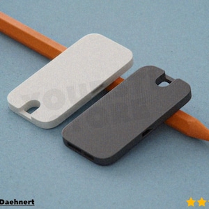 Może przedstawiać: Dwa prostokątne etui na telefon, jedno jasnoszare i jedno ciemnoszare, z zaokrąglonymi rogami i wycięciem na aparat. Pomiędzy nimi na niebieskiej powierzchni leży pomarańczowy ołówek.