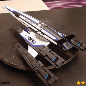 May include: A model of the Normandy SR2 spaceship, featuring a sleek design with white, black, and blue accents. The model has the text "NORMANDY" and "ASR2" printed on it. The spaceship is displayed on a dark circular base.