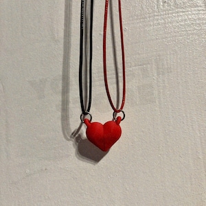 May include: A red heart pendant necklace with a black and red cord. The heart is connected to the cords with silver-colored rings. The necklace is displayed against a white wall.