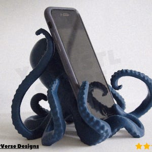 May include: A dark blue octopus phone stand supporting a black smartphone. The stand features multiple tentacles, designed to hold a phone upright. The product is set against a white backdrop. The brand name "MakerVerse Designs" is visible.