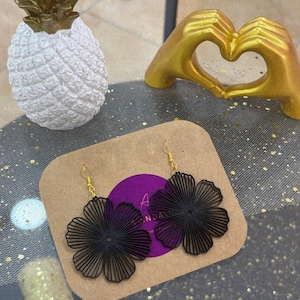 Puede incluir: Pendientes negros con forma de flor y ganchos dorados. Se exhiben sobre una tarjeta marrón con un círculo morado y texto. Una estatua de piña blanca y una estatua de manos doradas en el fondo.