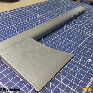 Puede incluir: Un hacha de metal gris con una cabeza rectangular y un mango largo. El mango tiene varias pequeñas hendiduras espaciadas uniformemente. El hacha está sobre una esterilla de corte azul con líneas de cuadrícula blancas. El texto "@Triple C Workshop" es visible.
