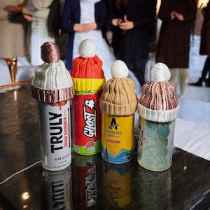 May include: Four beverage cans, each adorned with a miniature knitted hat. The cans include "Truly" and "Ghost" brands, with the hats in various colors like brown, white, and red. The cans are on a reflective surface.