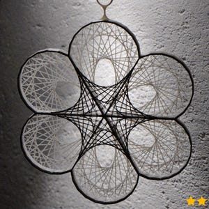 Puede incluir: Un colgante de pared geométrico con un diseño en forma de flor. La pieza presenta seis secciones circulares dispuestas alrededor de un elemento central en forma de estrella. Cada círculo está relleno de hilo blanco, y la estructura de conexión es negra.
