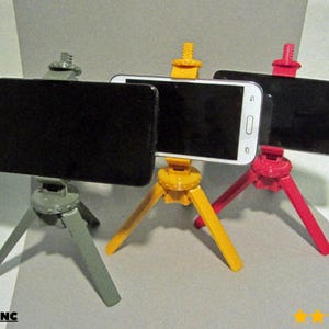 May include: Three brightly coloured smartphone tripods in grey, yellow, and red. Each tripod supports a smartphone, with the screens facing forward. The tripods have three legs and adjustable heads. The logo "@LoboCNC" is visible.