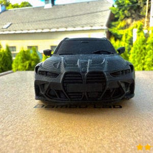 Op de afbeelding: Een zwart miniatuur automodel met gedetailleerde kenmerken, waaronder een grille, koplampen en zijspiegels. Het model staat op een lichtbruin oppervlak, met de tekst "Bambu Lab" zichtbaar. De achtergrond toont een huis en groen gebladerte.