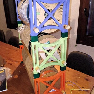 May include: A multi-colored, three-tiered spool rack. The rack is constructed from wood and painted in blue, green, and orange. Each level holds several spools of filament. The rack is designed to organize and display 3D printing filament.