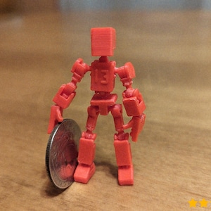 Puede incluir: Una pequeña figura roja articulada, sosteniendo una moneda. La figura tiene una cabeza y un cuerpo en forma de bloque, con brazos y piernas articulados. El número "13" es visible en el torso. La figura mide unos 5 cm de altura.
