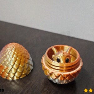 Puede incluir: Un huevo de dos partes impreso en 3D con un dragón dentro. El huevo está abierto, revelando una figura de dragón. El huevo y el dragón son en tonos dorados, naranjas y marrones. El dragón tiene ojos negros.