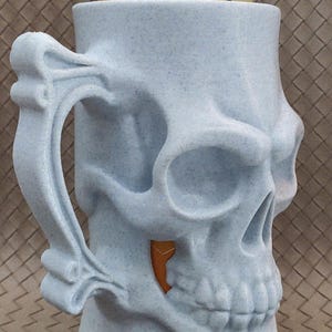 May include: A light blue skull-shaped beer stein with a handle. The stein is designed to hold a can, visible at the top. The design includes detailed facial features and a textured surface. The handle is intricately shaped.