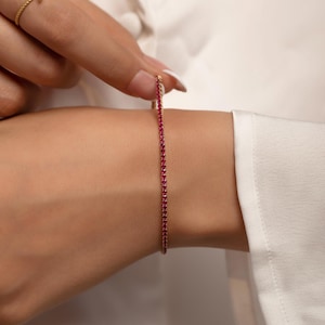 Puede incluir: Un brazalete dorado con una línea de pequeñas piedras preciosas rojas. También se ve un anillo dorado a juego con un diseño floral. La joyería se presenta sobre un fondo blanco, lo que sugiere un enfoque en los accesorios.