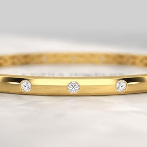 Peut inclure: Un bracelet jonc en or avec trois pierres précieuses rondes et transparentes espacées sur le devant. Le bracelet a une finition lisse et polie et un motif géométrique subtil. Le fond est blanc et flou.