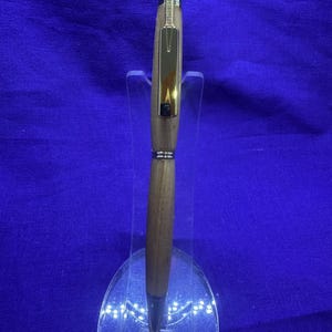 May include: A wooden pen with a gold-colored clip and accents. The pen has a light brown barrel and a silver-colored tip. The pen is displayed on a clear acrylic stand against a blue background.
