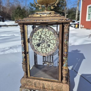 Peut inclure: Une horloge ancienne en laiton et verre au design orné. L'horloge présente un cadran de couleur claire avec des chiffres noirs et des aiguilles décoratives. Le cadre de l'horloge est orné d'accents d'émail colorés et d'un dessus décoratif.