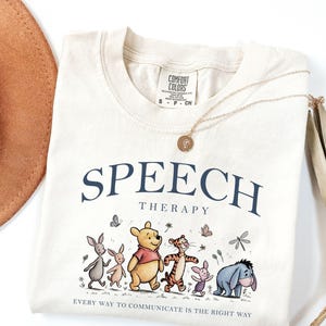 May include: A cream-colored t-shirt featuring the text "SPEECH THERAPY" in dark blue, followed by a graphic of Winnie the Pooh and friends. Below the characters, the text reads "EVERY WAY TO COMMUNICATE IS THE RIGHT WAY." The shirt has a Comfort Colors label.