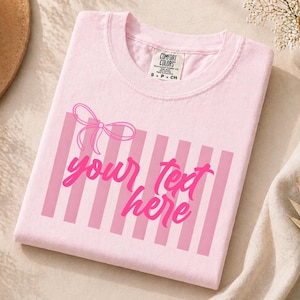 May include: A light pink t-shirt with a pink bow and vertical stripes design. The text "your text here" is written in a cursive font. The shirt has a Comfort Colors tag.