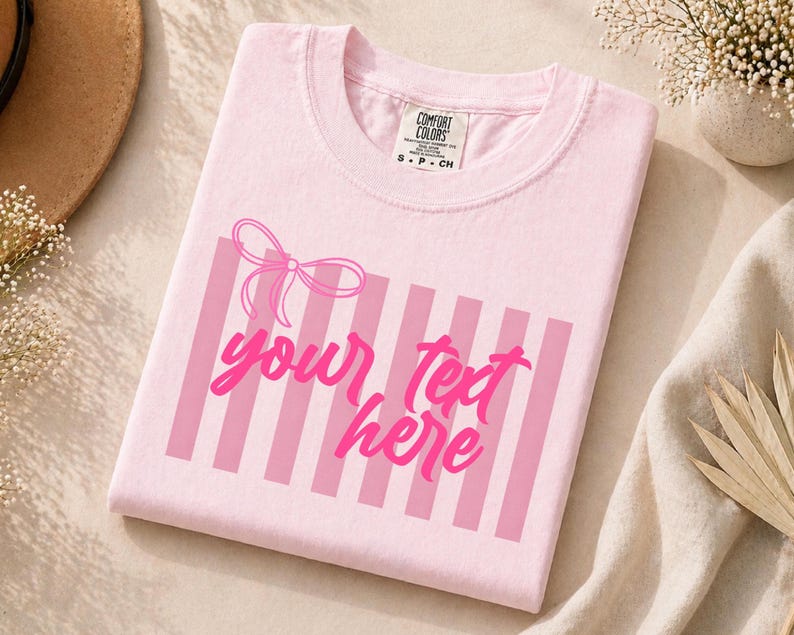 May include: A light pink t-shirt with a pink bow and vertical stripes design. The text "your text here" is written in a cursive font. The shirt has a Comfort Colors tag.