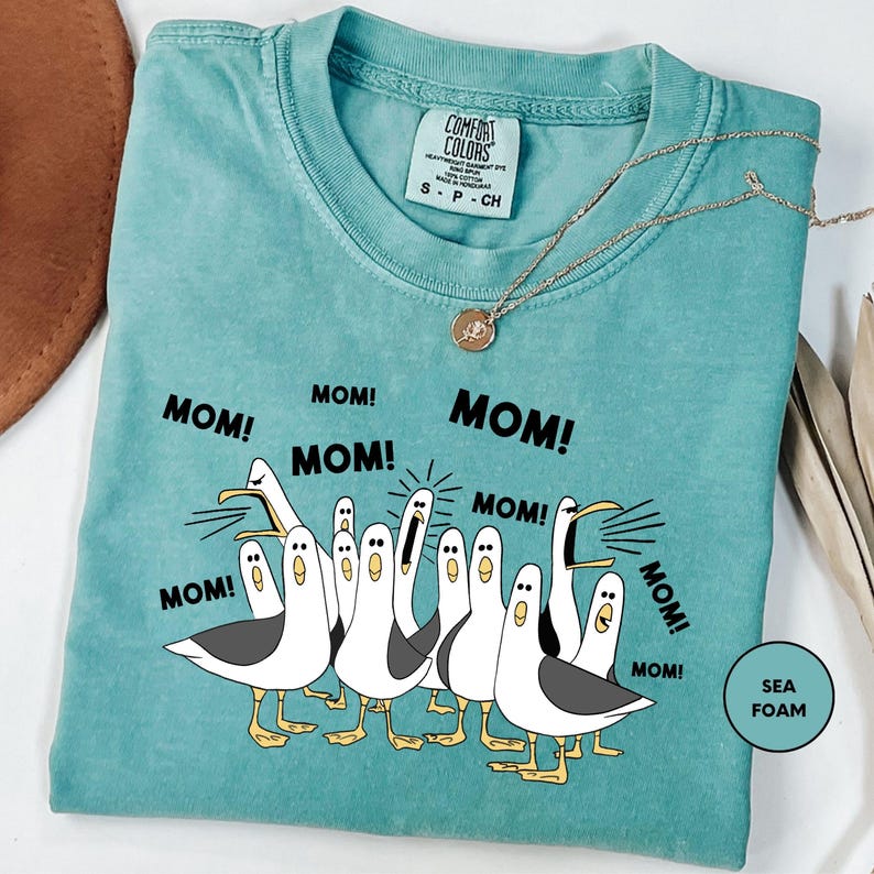 A turquoise t-shirt with a graphic design of a group of seagulls and the text "MOM! MOM! MOM! MOM!" printed on it.